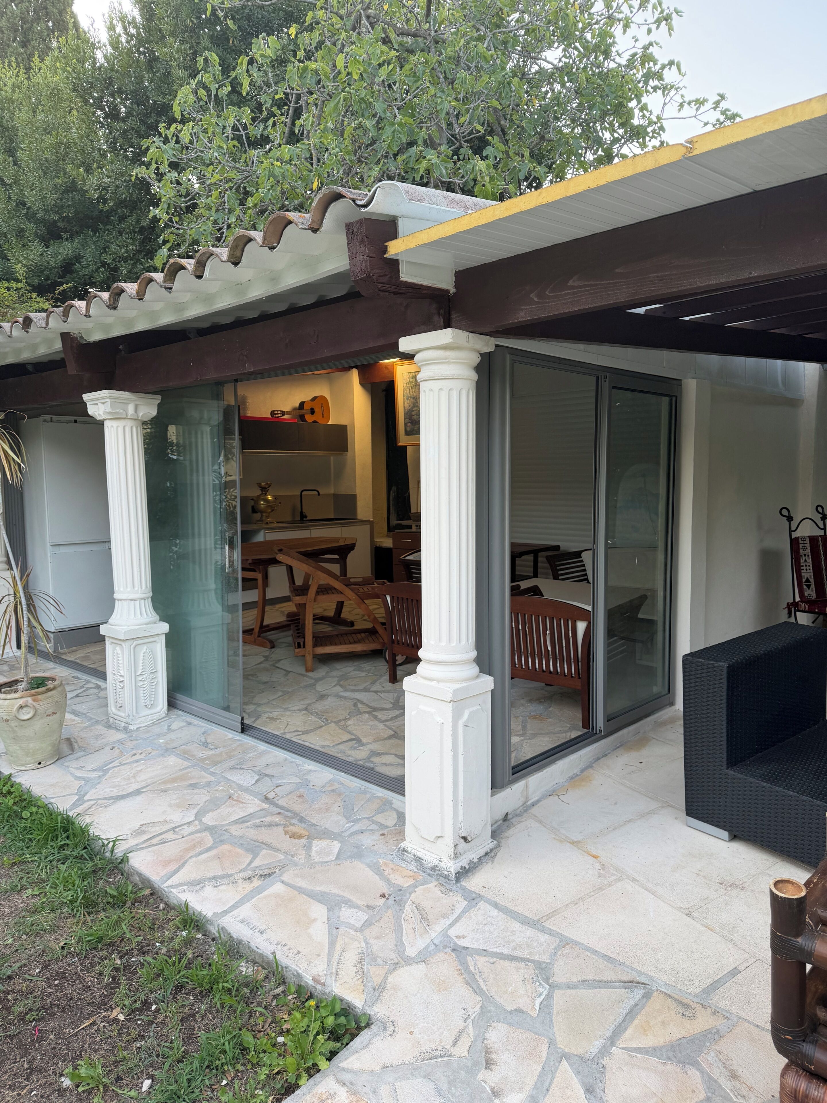 Une terrasse couverte avec un sol en pierre, des colonnes blanches et une porte vitrée donnant sur un espace intérieur comprenant une table et des chaises.