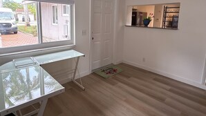 A room with a glass table, a white door, a window with a view of parked cars, and a small kitchen area with a sink and a plant. - Sunny Cove Retreat: Walk to Beach, Dining & Marina (Deerfield Beach)