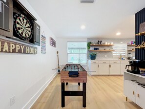 Ein Zimmer mit Tischfußball, Dartscheibe und Küchenzeile mit Regalen und Geräten.