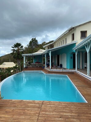 Piscina com deck de madeira, área de varanda coberta e um edifício com várias janelas e varanda.