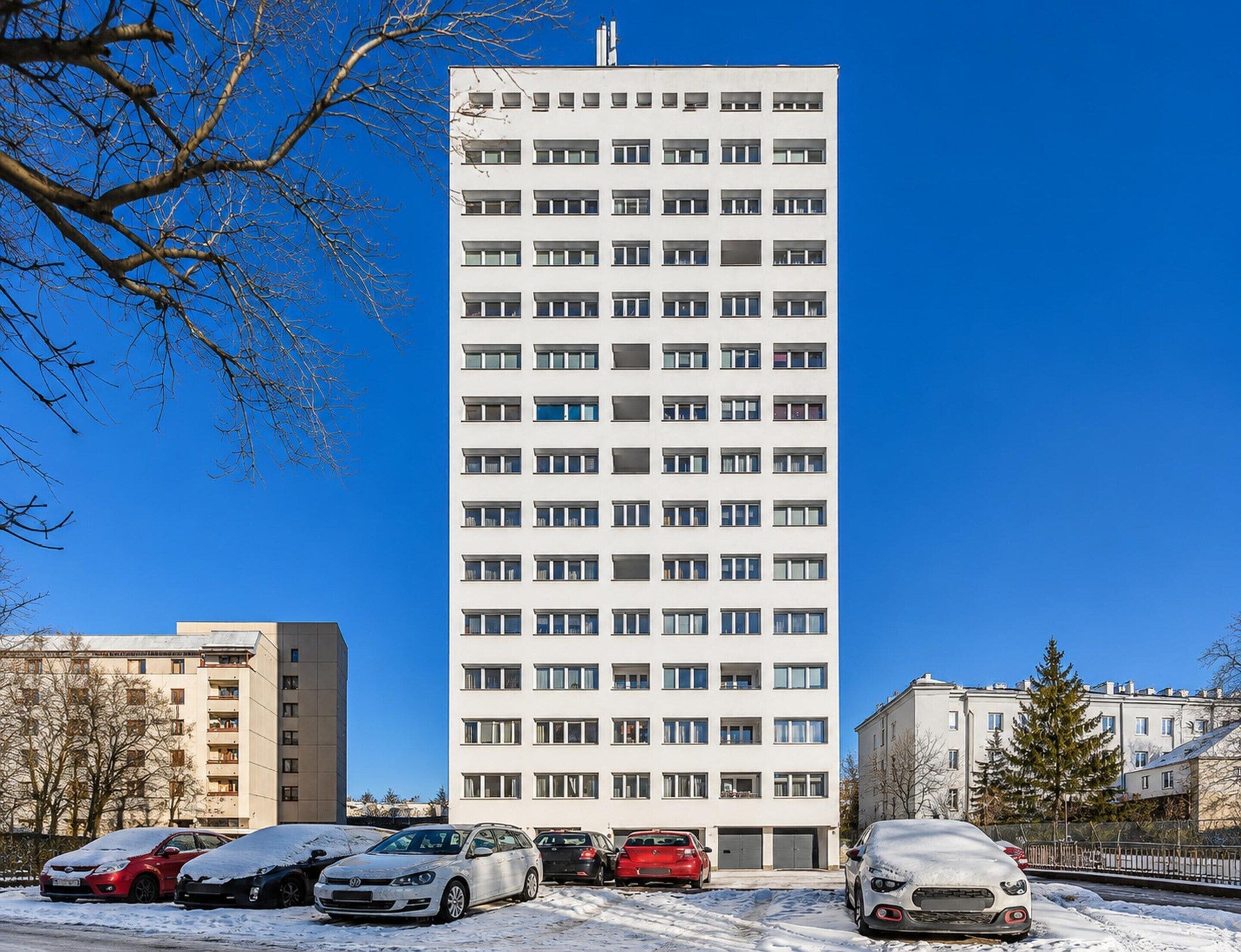 A multi-story residential building with a uniform facade, several balconies, and a parking area with cars.