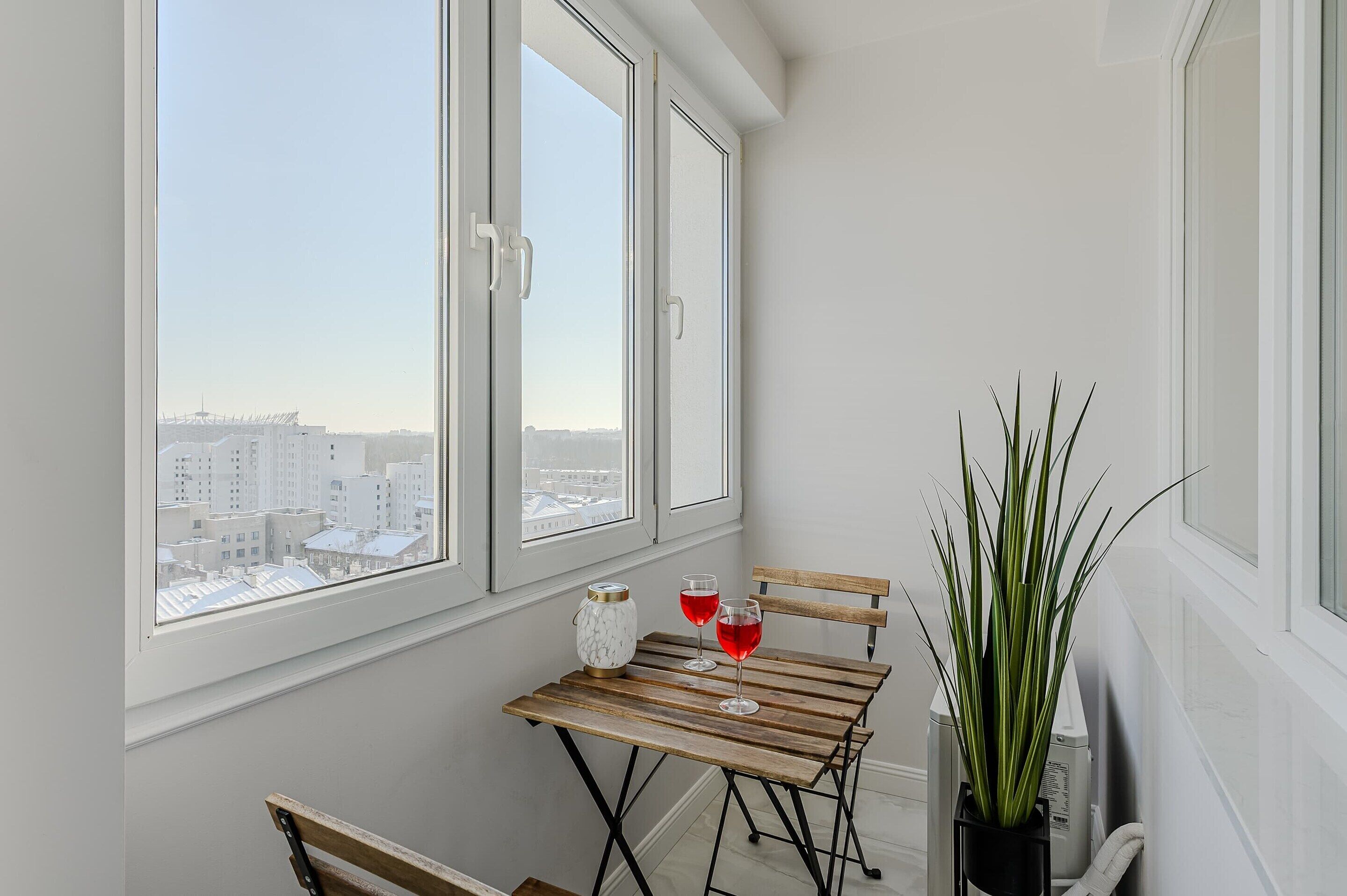 A balcony with a view of a cityscape, a wooden table with two glasses of red liquid, and a potted plant.
