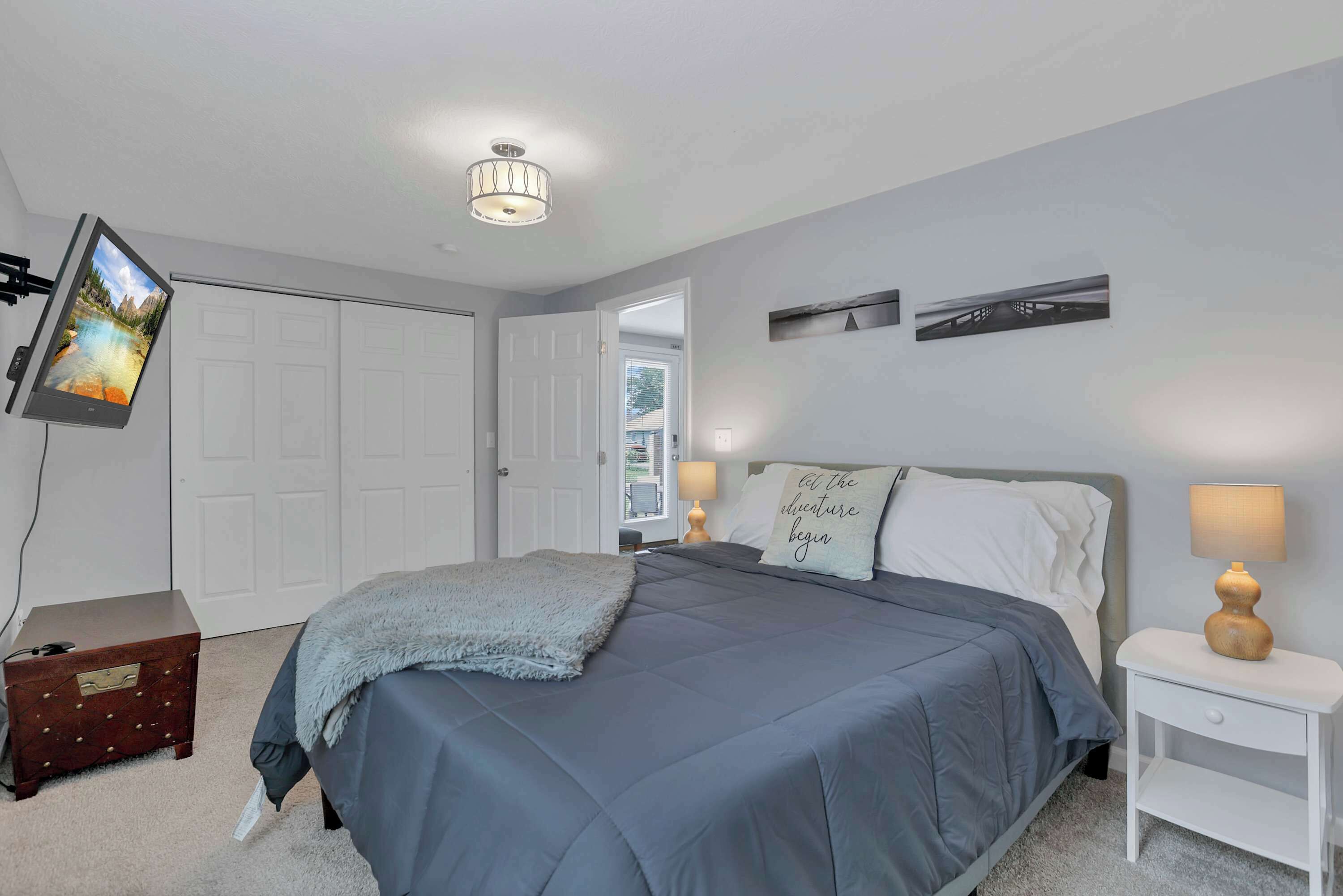 A bedroom with a bed, bedside tables, a desk, a chair, a TV mounted on the wall, and a door.