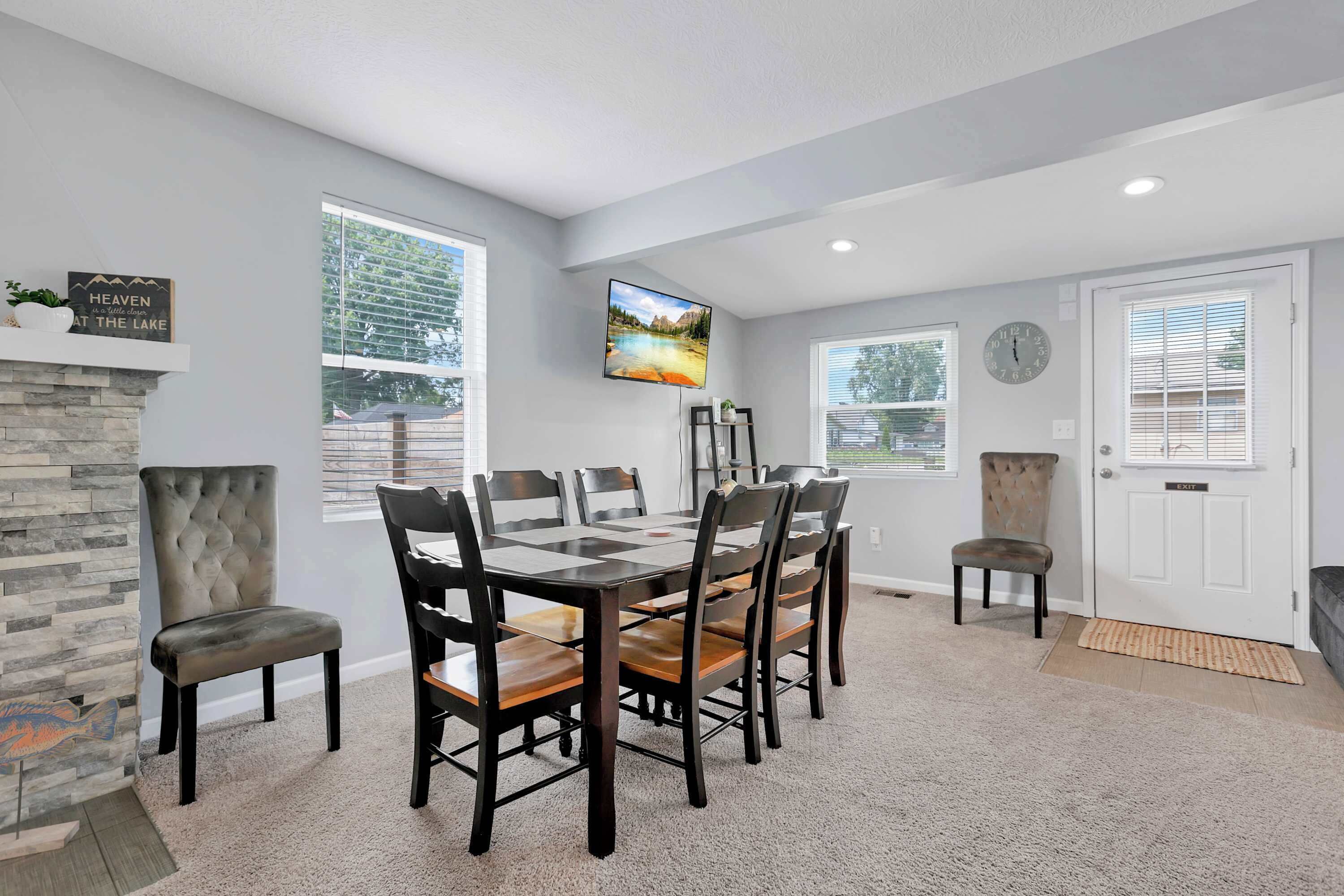 A dining area with a stone fireplace, a large window with blinds, a flat-screen TV mounted on the wall, and a clock on the wall.