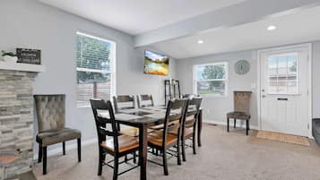 A dining area with a stone fireplace, a large window with blinds, a flat-screen TV mounted on the wall, and a clock on the wall.