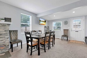 A dining area with a stone fireplace, a large window with blinds, a flat-screen TV mounted on the wall, and a clock on the wall.