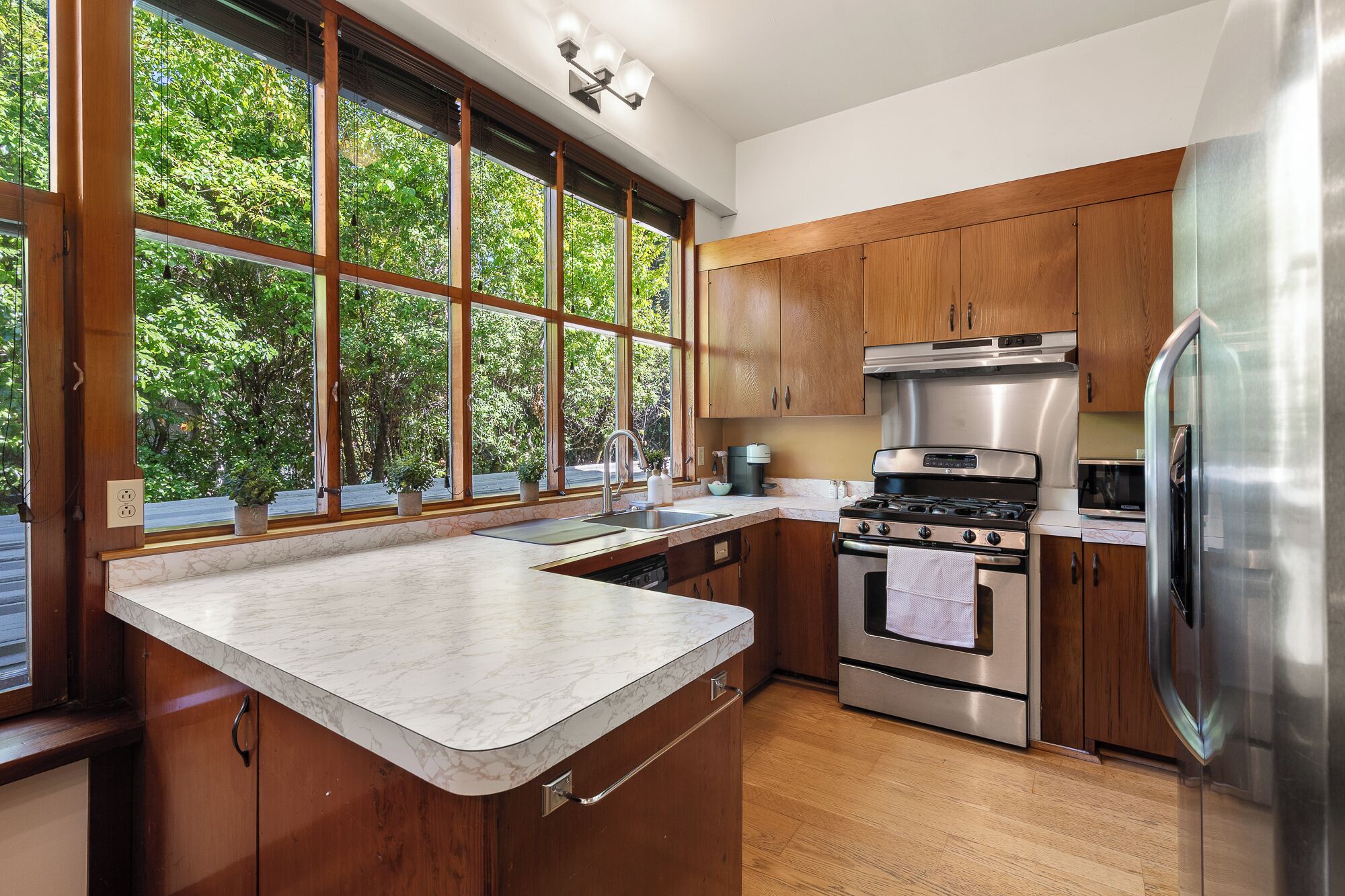 A modern kitchen with wooden cabinets, stainless steel appliances, and a large window overlooking greenery.