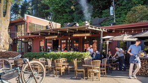 An outdoor seating area with wooden chairs and tables, a bicycle, and a building with large windows and a chimney emitting smoke.