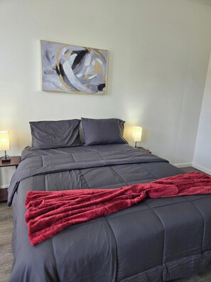 A bedroom with a bed, two bedside lamps, a red blanket, and a wall-mounted abstract painting.