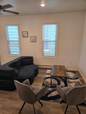 A living room with a sectional sofa, two armchairs, a coffee table, and a rug. There are two windows with blinds and a ceiling fan.