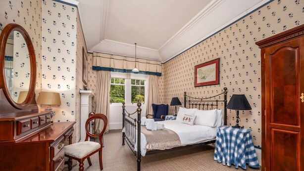 A bedroom with a bed, a chair, a dresser, a mirror, and a window with curtains. - Dunkeld House - Luxury Cottages (Dunkeld)
