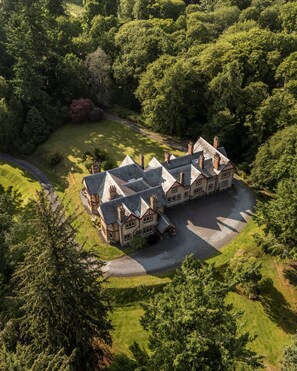 A large, multi-story building with a symmetrical roofline and multiple chimneys, surrounded by lush greenery and a well-maintained lawn. - Dunkeld House - Luxury Cottages (Dunkeld)