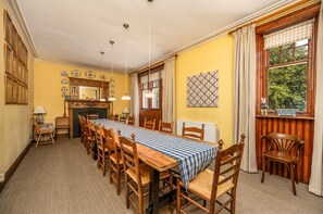A dining room with a long wooden table, chairs, a fireplace, and a view of greenery through the window. - Dunkeld House - Luxury Cottages (Dunkeld)