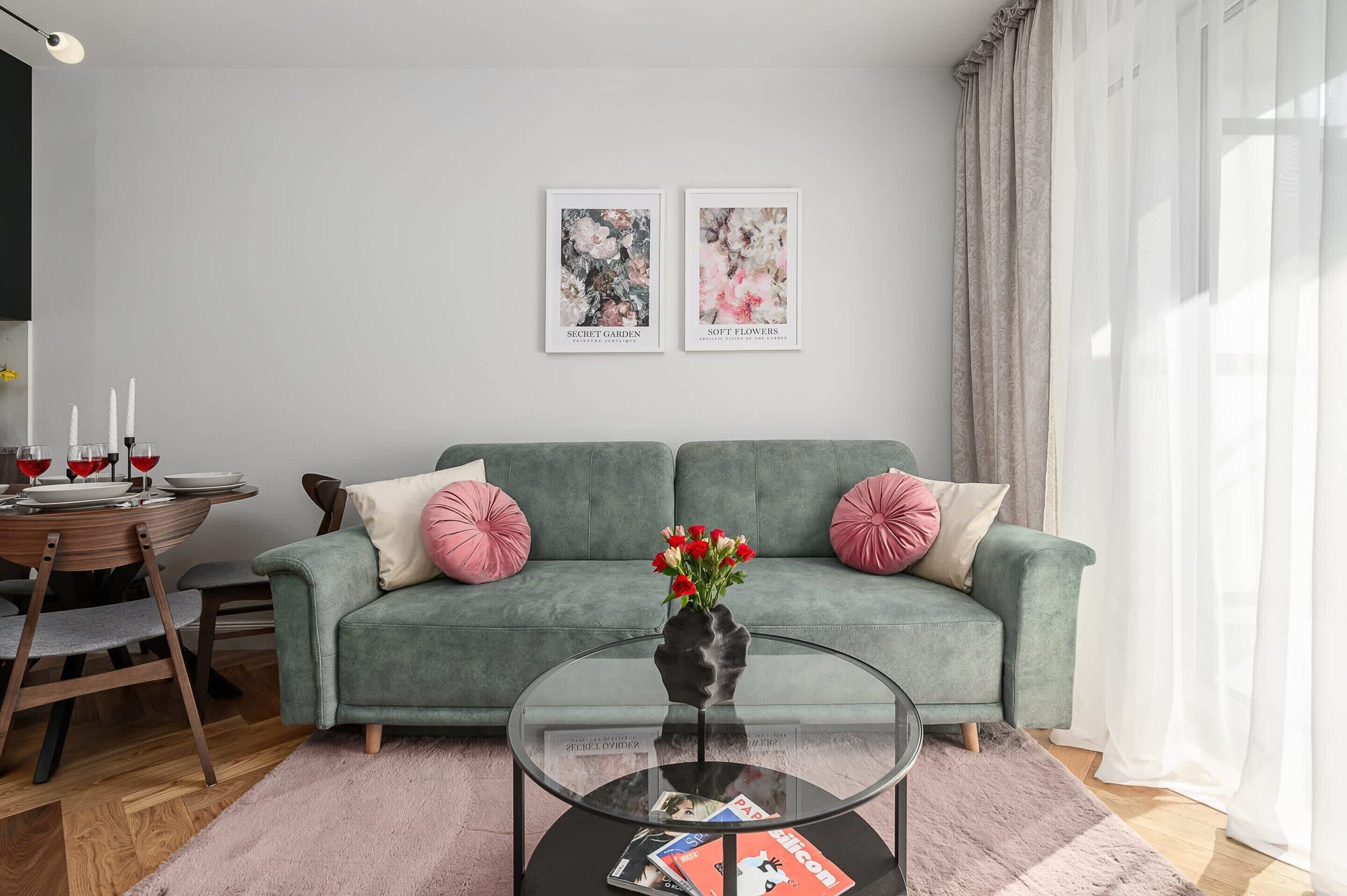 A modern living room with a grey sofa, two pink cushions, a glass coffee table with magazines, and a vase with red flowers.