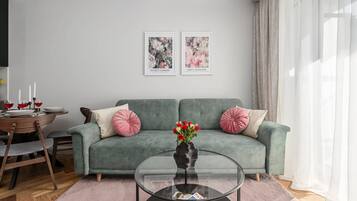 A modern living room with a grey sofa, two pink cushions, a glass coffee table with magazines, and a vase with red flowers.