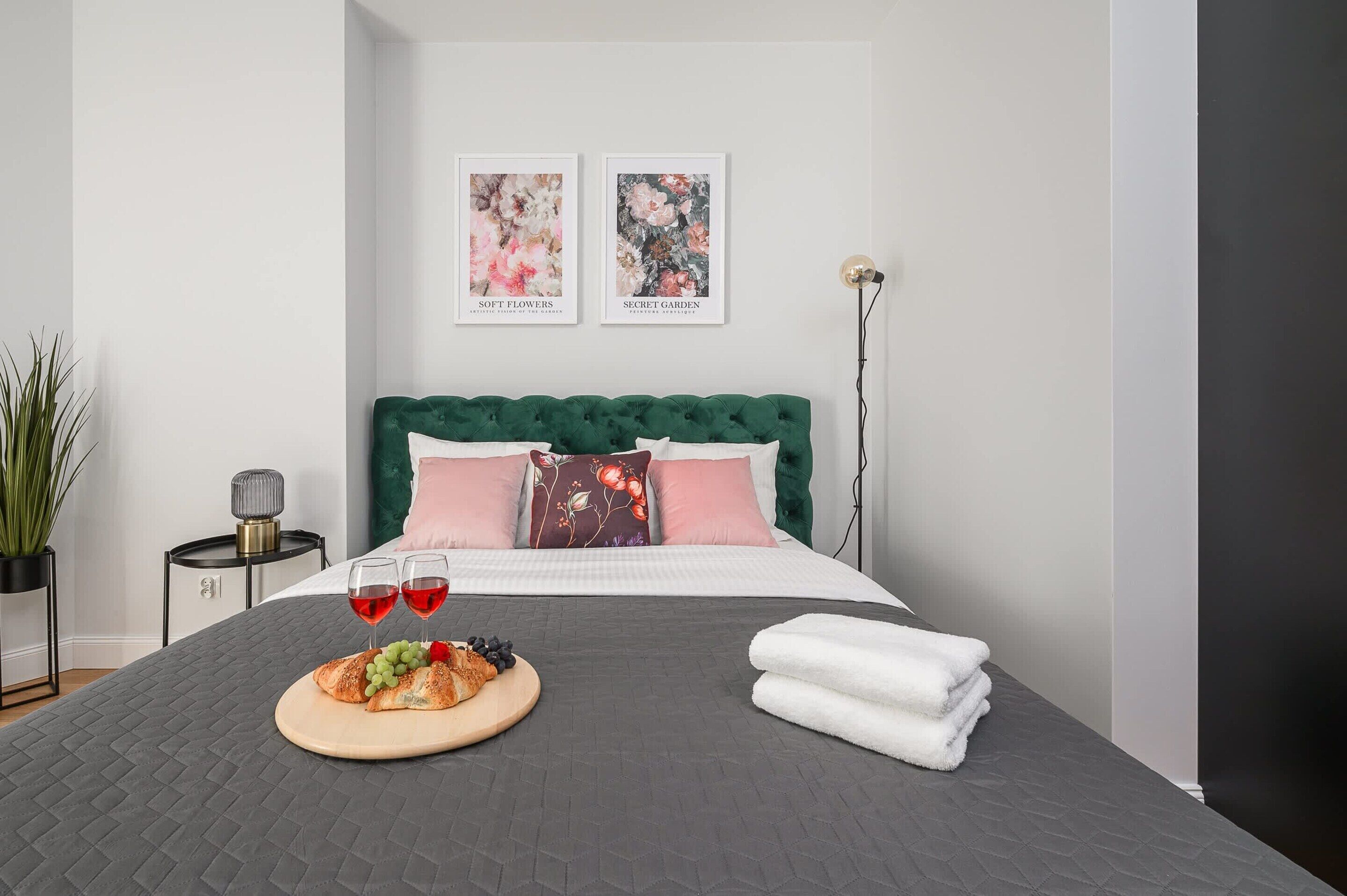 A neatly arranged bedroom with a green upholstered headboard, a bed with pink and white pillows, two framed artworks on the wall, a bedside table with a lamp, and a breakfast setup on the bed.