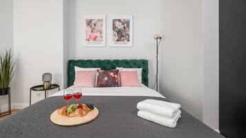 A neatly arranged bedroom with a green upholstered headboard, a bed with pink and white pillows, two framed artworks on the wall, a bedside table with a lamp, and a breakfast setup on the bed.