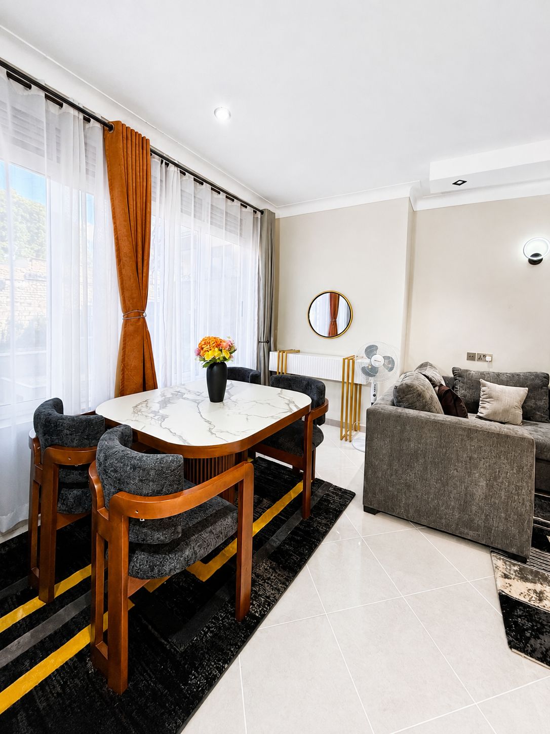 Uma sala de estar moderna com área de jantar, apresentando uma mesa de mármore, cadeiras estofadas cinzentas, um sofá cinza e um tapete preto e amarelo.