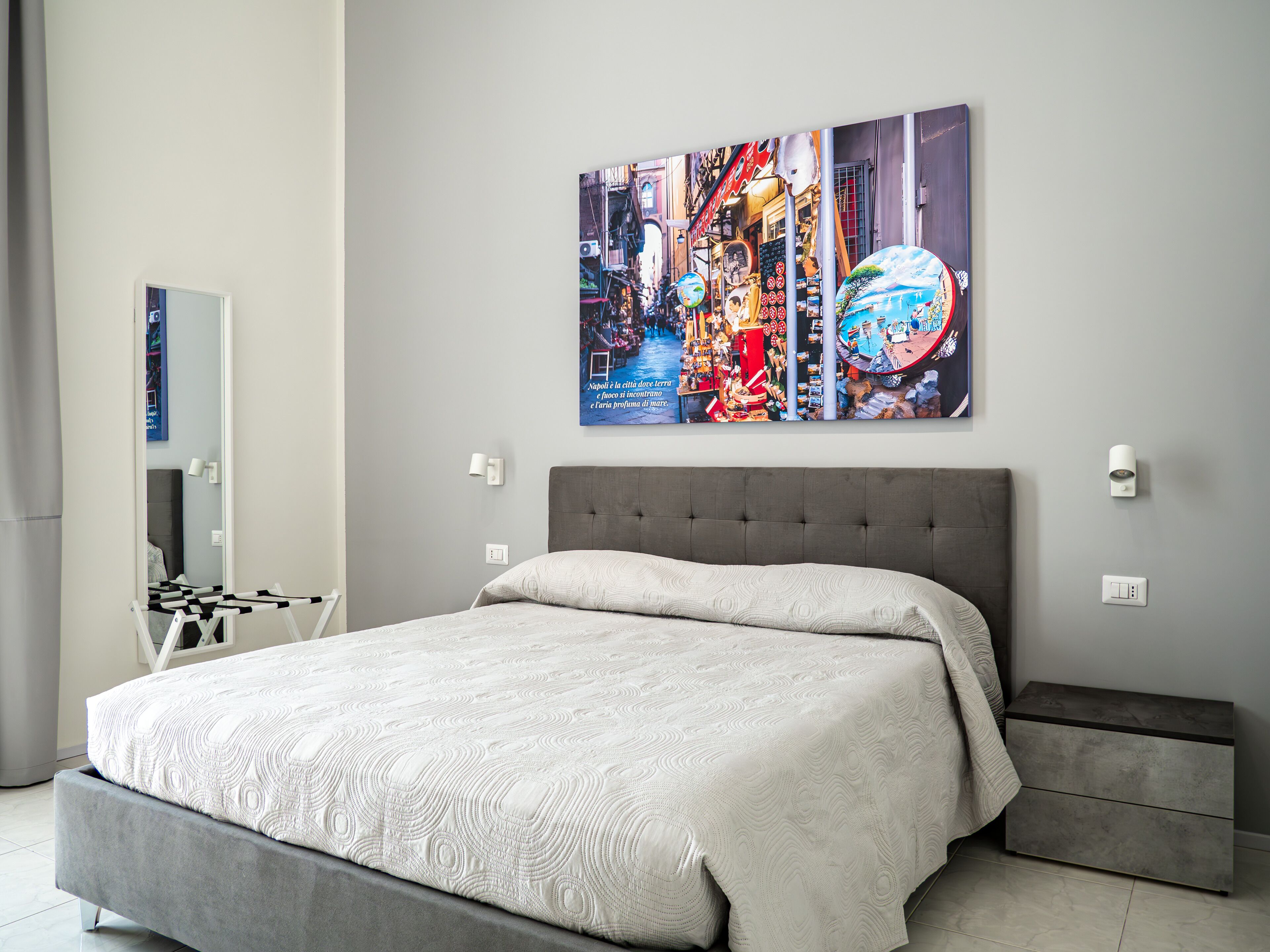 Ein modernes Schlafzimmer mit einem grau gepolsterten Bett, einer grauen Bank und einem Wandbild mit einer lebendigen Stadtszene.