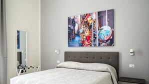 Et moderne soveværelse med en grå polstret seng, en grå bænk og et vægmaleri der viser et livligt bybillede.