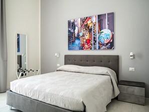 A modern bedroom with a grey upholstered bed, a grey bench, and a wall-mounted artwork depicting a vibrant city scene. - Vesuvian Tales (San Giorgio a Cremano)