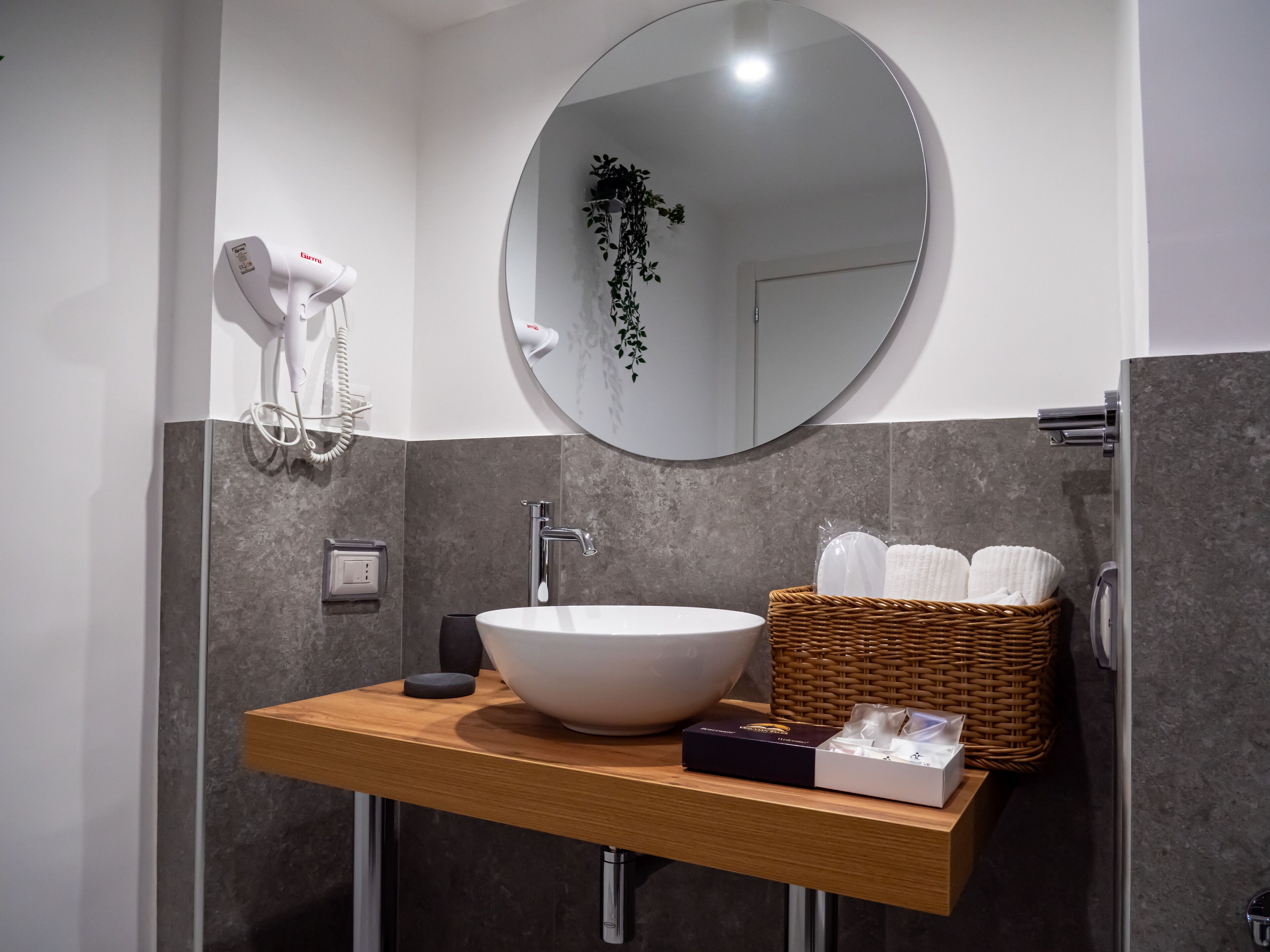 Een moderne badkamer met een ronde spiegel, een witte wasbak, een haardroger, een mandje met toiletartikelen en een kleine plant.
