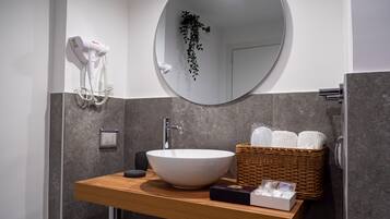 Een moderne badkamer met een ronde spiegel, een witte wasbak, een haardroger, een mandje met toiletartikelen en een kleine plant.