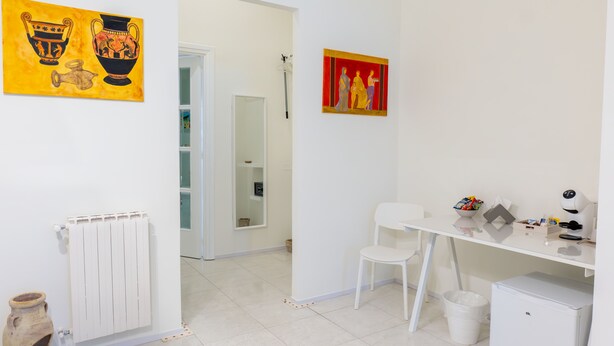 Et lille, rent værelse med en hvid radiator, en hvid stol, et lille hvidt bord og et hvidt skab. Der hænger to billeder på væggen.