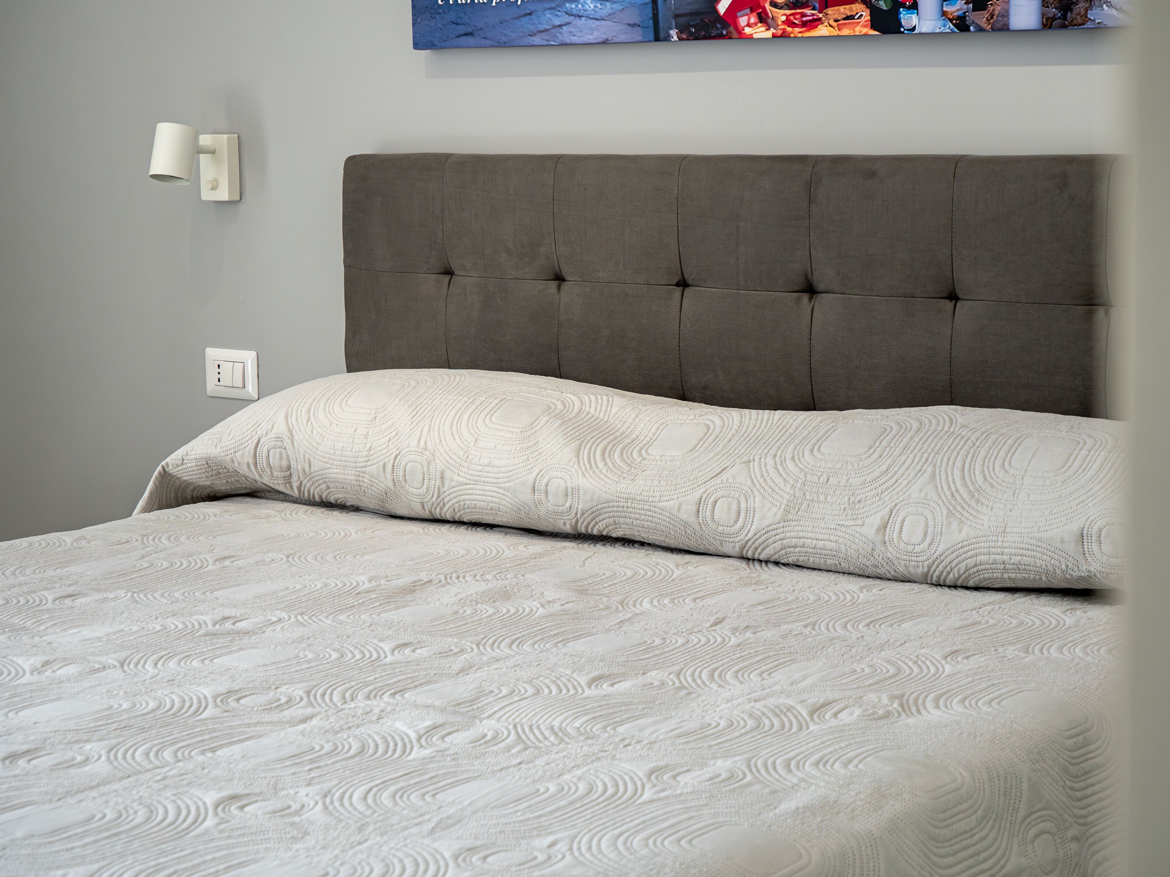 Una cama con cabecera capitonada, colcha estampada y lámpara fijada a la pared.
