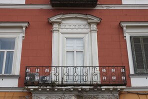 Fachada de edificio con una puerta blanca central, flanqueada por ventanas con contraventanas y barandilla de balcón decorativa.