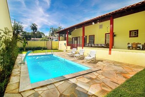 A swimming pool with a stone patio, surrounded by a yellow building with red pillars and a tiled roof. - Casa Del Rey (Pirenópolis)