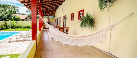 Un espacio exterior techado con una hamaca, sillas junto a la piscina y piso de baldosas.