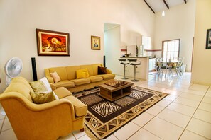 A living room with a sofa, a coffee table, and a fan. There is a kitchen area with a dining table and chairs visible in the background. - Casa Del Rey (Pirenópolis)
