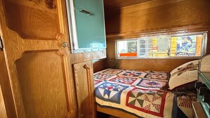 Ein kompaktes Wohnmobil-Interieur mit Holzboden, einem Bett mit Quiltdecke, einer Küchenzeile und einem Fenster mit Außenblick.