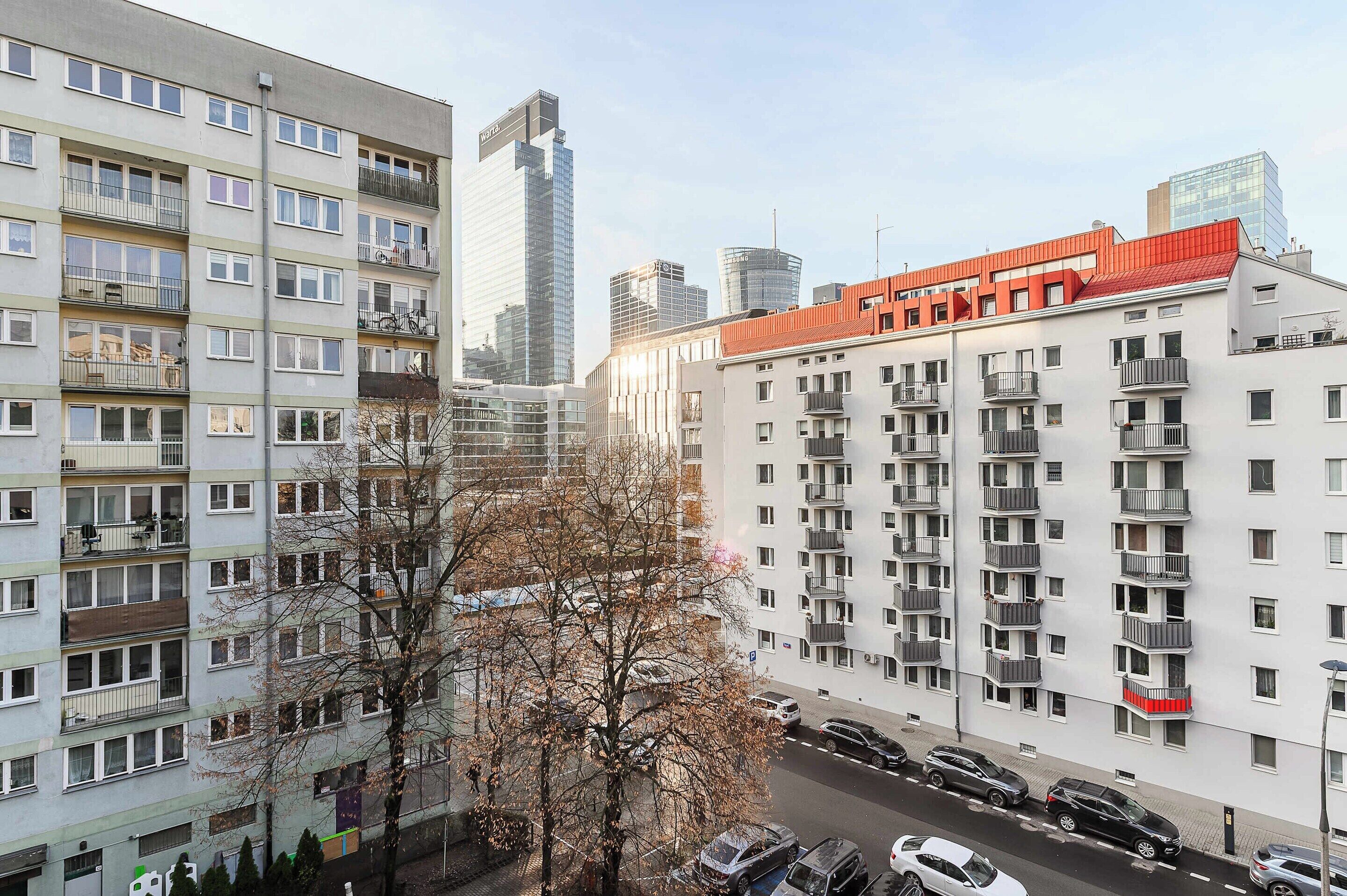 Um edifício residencial com vários andares, varandas, área de estacionamento e edifícios altos modernos ao fundo.