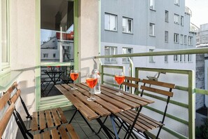 Ein Balkon mit Holztischen und -stühlen, zwei Gläsern Rotwein und Blick auf ein Gebäude mit zahlreichen Fenstern.