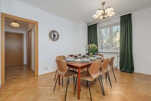 A dining area with a wooden table set for four, surrounded by six chairs, a wall clock, and a window with curtains. - Stylish Warsaw Apartment With Balocny (Warsaw)