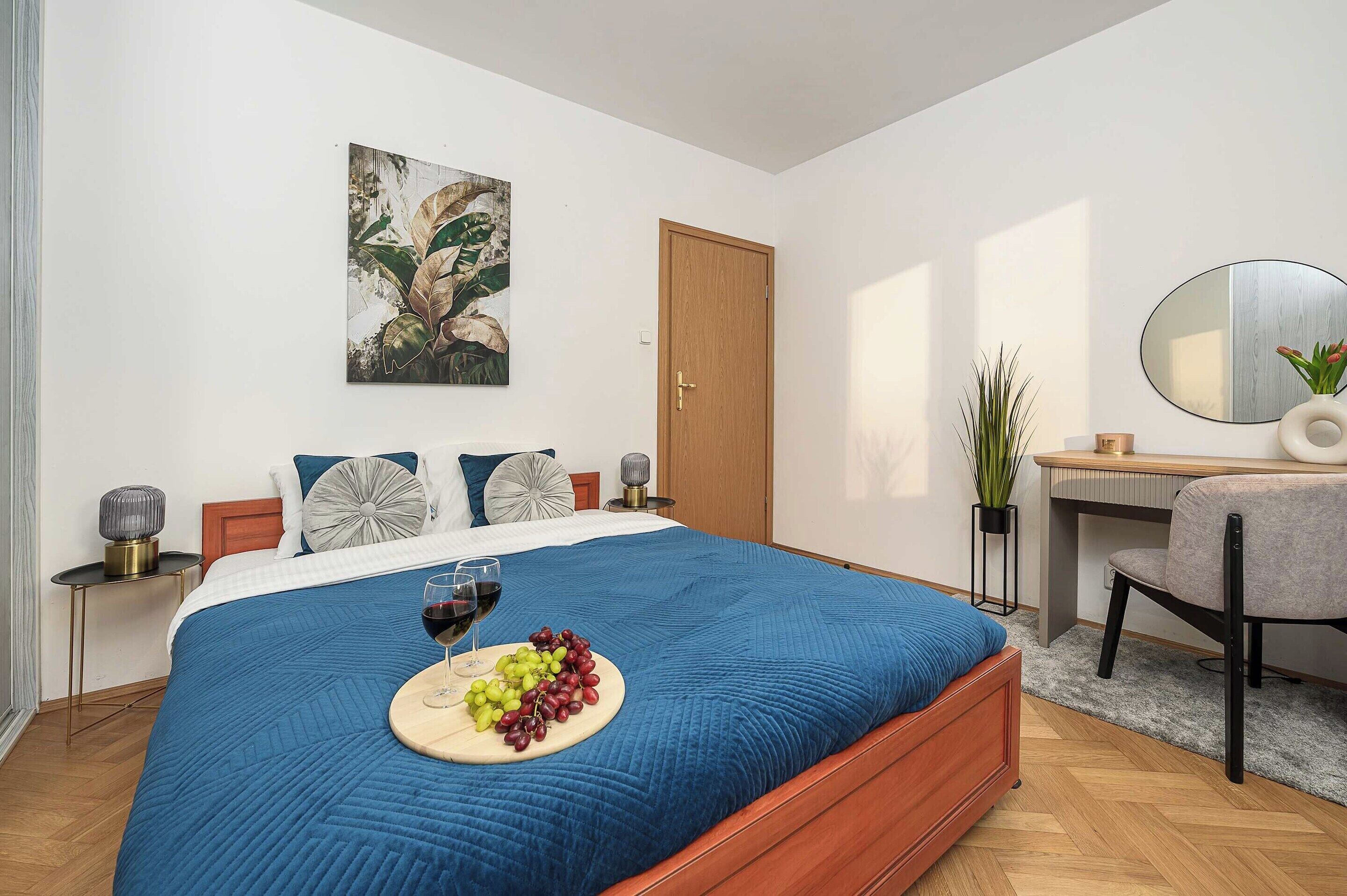 A bedroom with a bed, a bedside table, a desk, a chair, a mirror, and a wall with a painting.