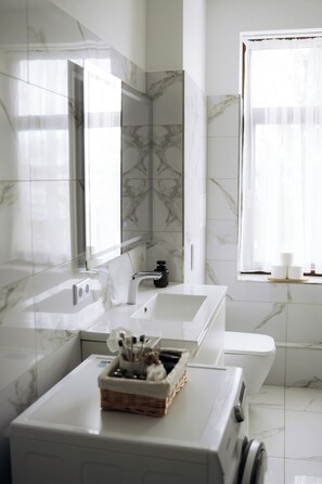 Een moderne badkamer met een marmeren spatwand, een grote spiegel, een witte wastafel, een toilet en een raam met dunne gordijnen.