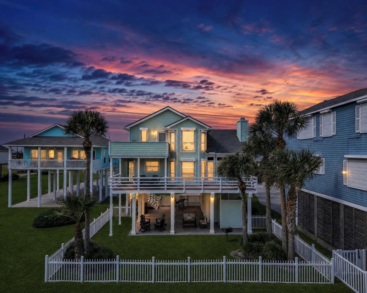 A two-story house with a covered patio, a fenced yard, and a view of the sunset.