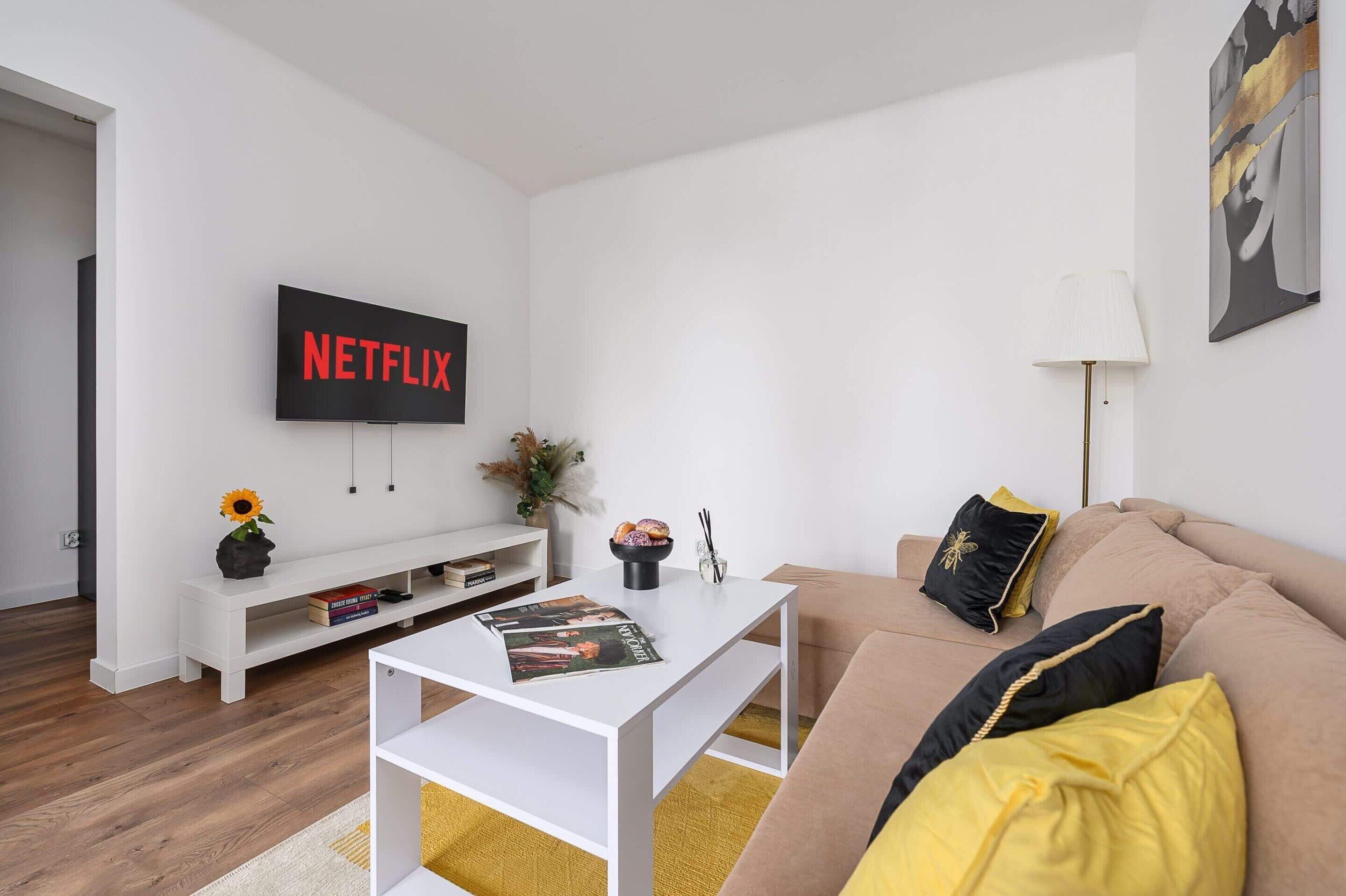 Ett modernt vardagsrum med en platt-TV som visar ”NETFLIX”, ett vitt soffbord, en beige soffa med gula kuddar och ett vitt sidobord med en vas och böcker.
