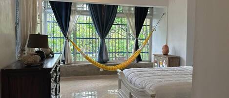 A bedroom with a bed, a nightstand, a lamp, a window with curtains, and a hammock.