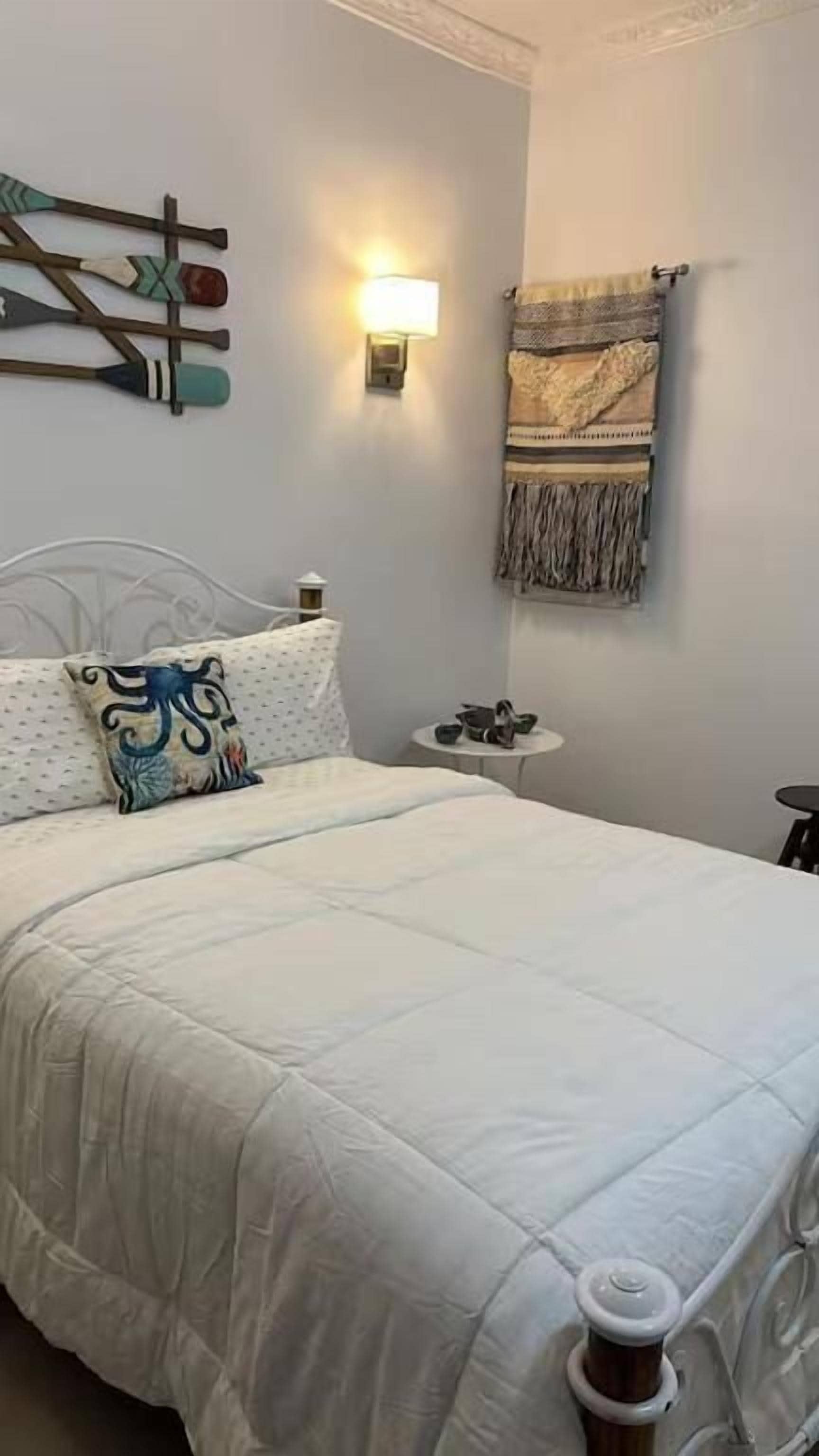 Quarto com uma cama, uma luminária na parede, uma pequena mesa redonda com abajur e um ornamento decorativo na parede.