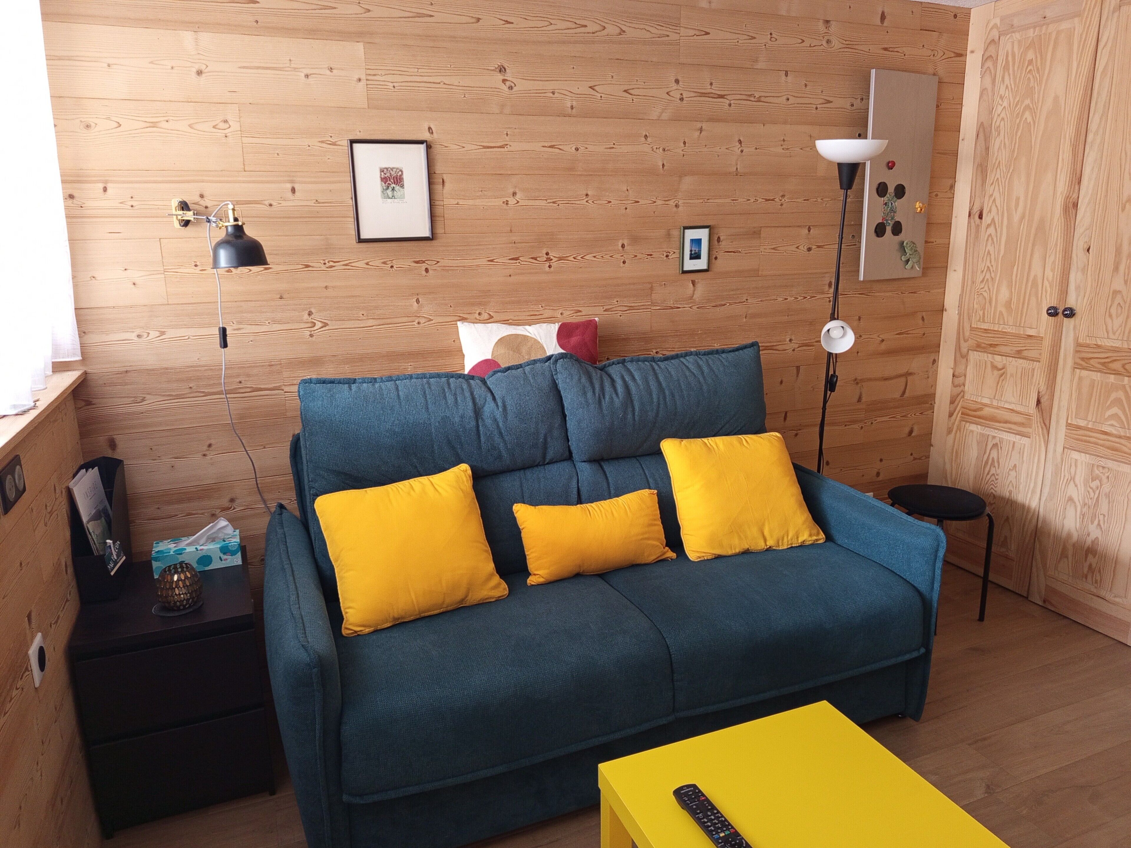 Ein Zimmer mit einem blauen Sofa, gelben Kissen, einem kleinen Tisch und einer Lampe.