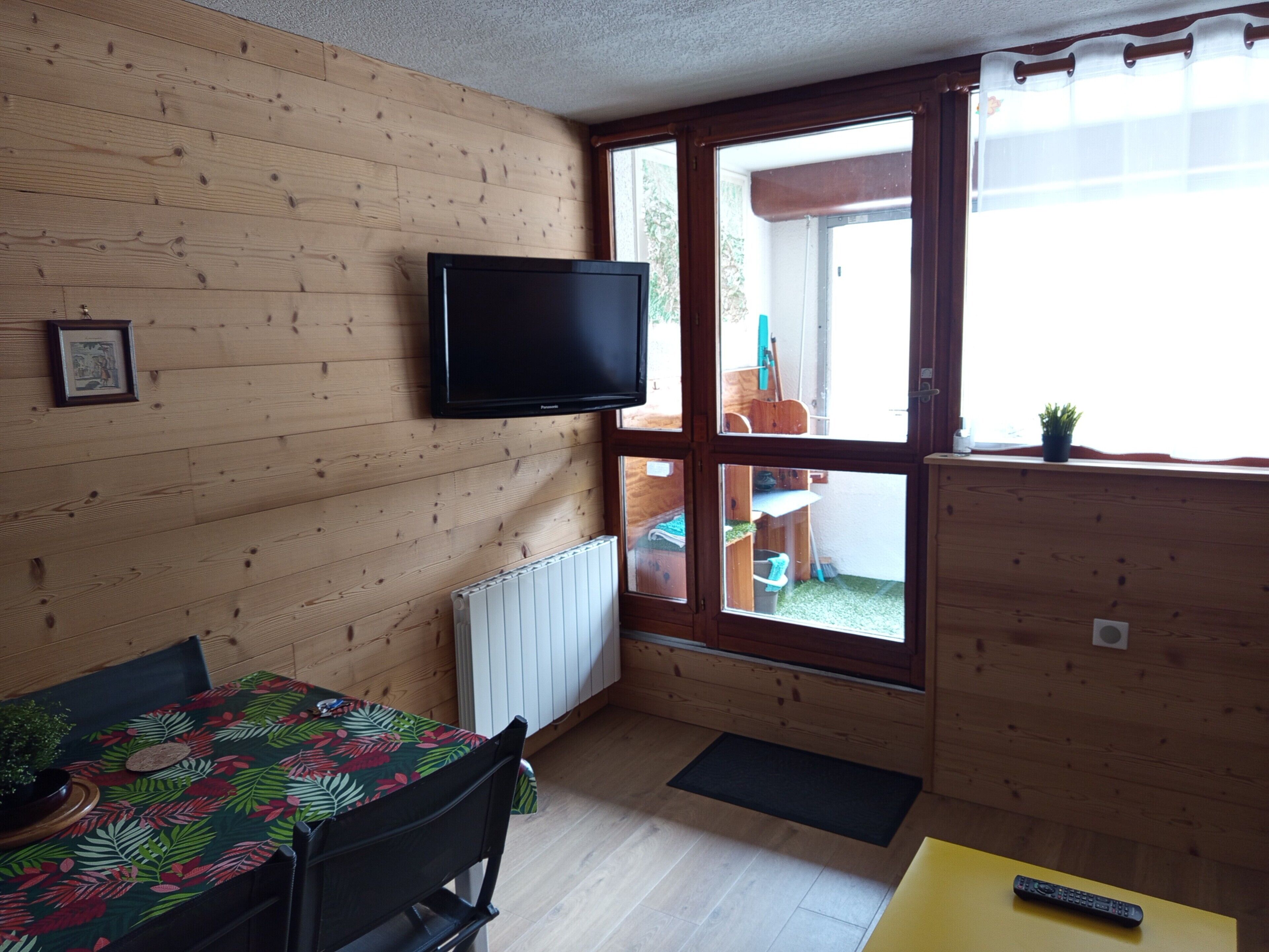 A room with a wooden interior, a television mounted on the wall, a dining table with chairs, and a window with a view of the outside.