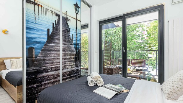 Ein Schlafzimmer mit einem großen Bett, Blick auf einen Steg durch eine Glastür und einem Balkon mit Gartenmöbeln.