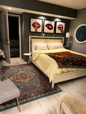 Een slaapkamer met een bed, nachtkastje, lamp, deur en een muur met kunst.