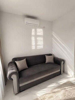 Ein Eckzimmer mit einer grauen Couch, einer weißen Klimaanlage und einem Fenster, das Schatten an die Wand wirft.