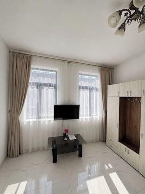 Ein Zimmer mit einem wandmontierten Flachbildfernseher, einem Holzschrank, einem kleinen Tisch mit einer Vase und einem Fenster mit durchsichtigen Vorhängen.