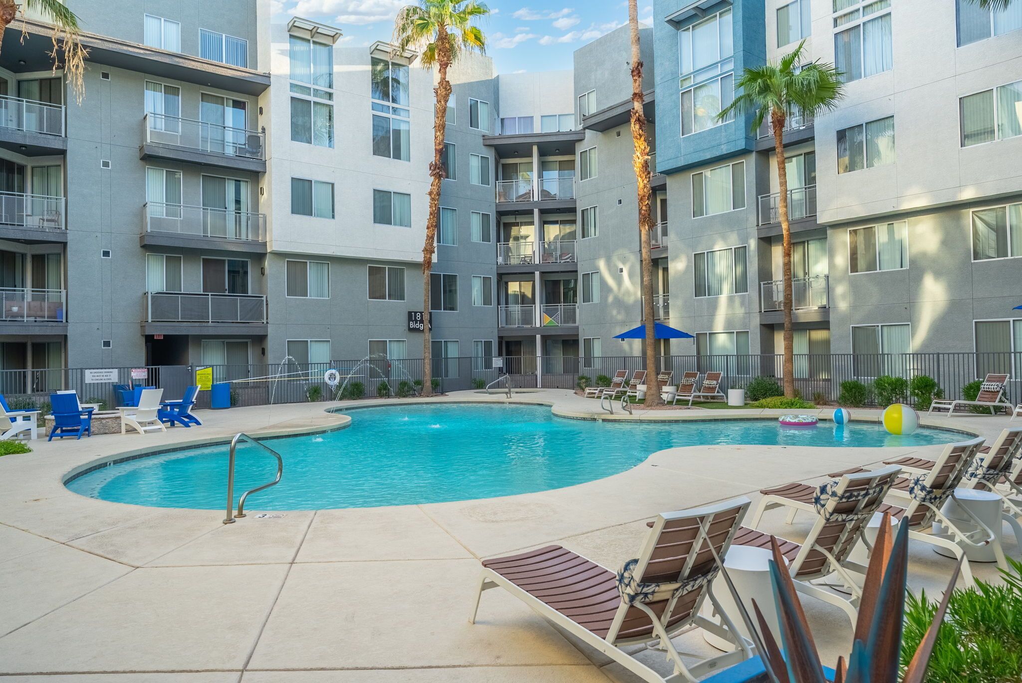 A modern apartment building with a swimming pool, lounge chairs, and palm trees.